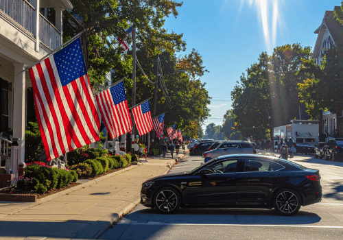 Black car parked on a sunny street lined with American flags, trees, and homes during Memorial Day.