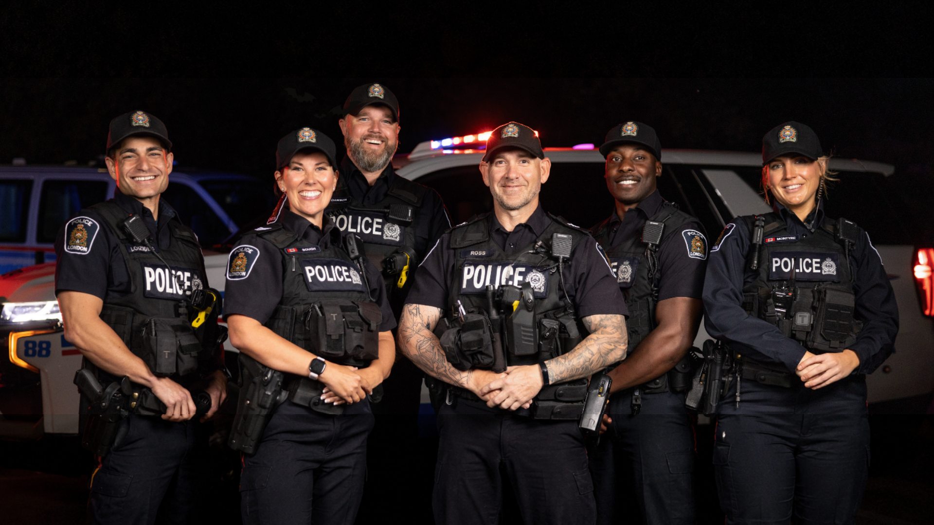 Six policiers en uniforme sombre se tiennent ensemble la nuit, souriant devant un véhicule.