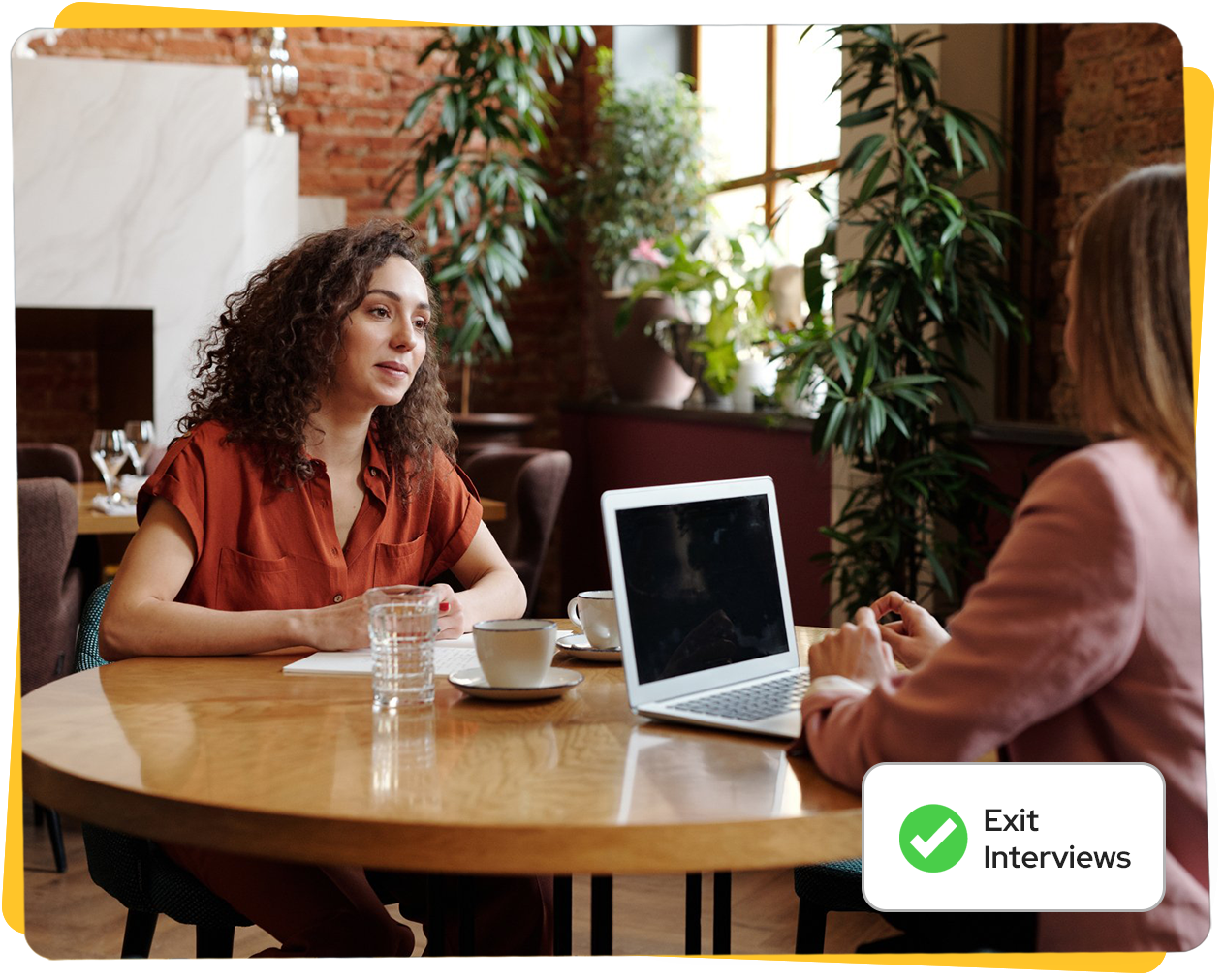 Two women at a table in a cafe, one speaking, laptop present,