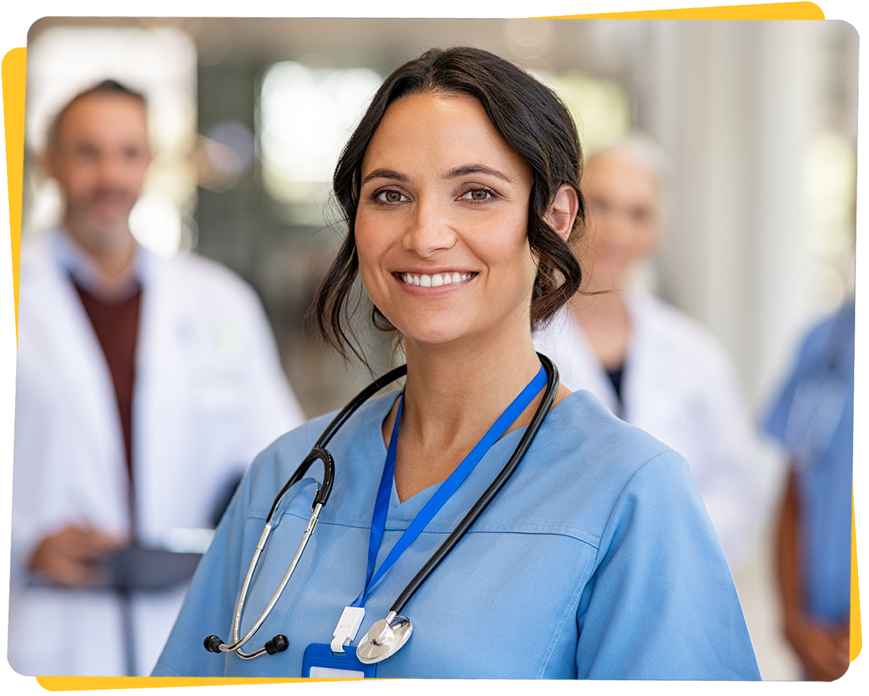 Nurse in blue scrubs with stethoscope, smiling, in a medical setting, with colleagues in the background.