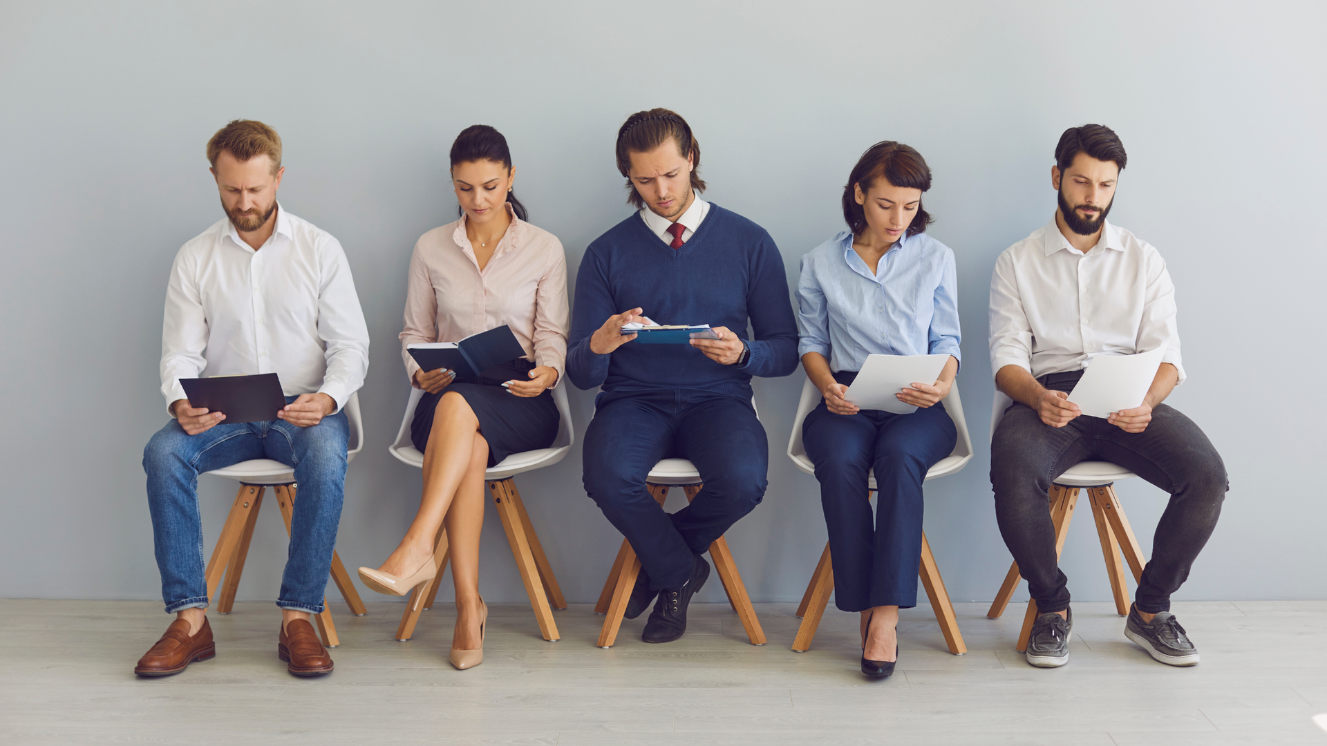 Five people seated in a row on chairs, each looking down at a clipboard in a waiting room.