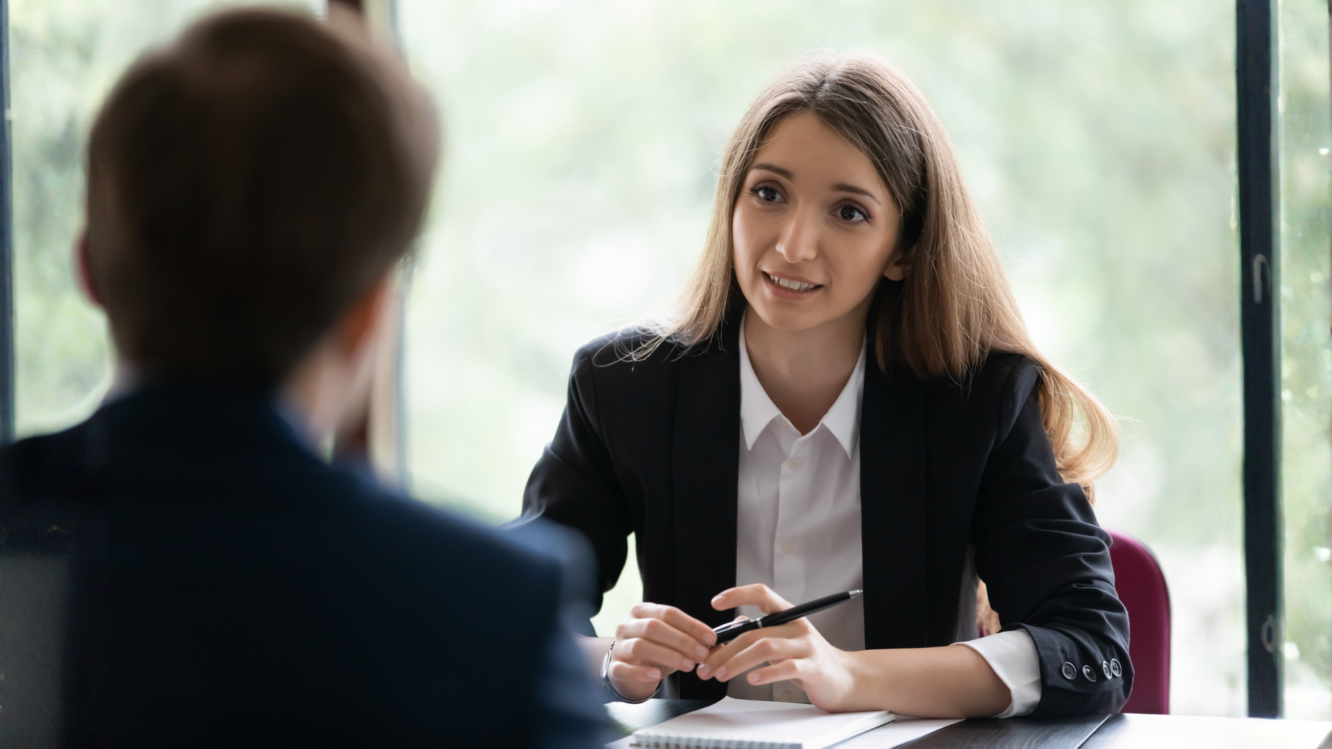 Woman in black blazer at a table interviewing a person. She holds a pen, looking and speaking. Window in the background.