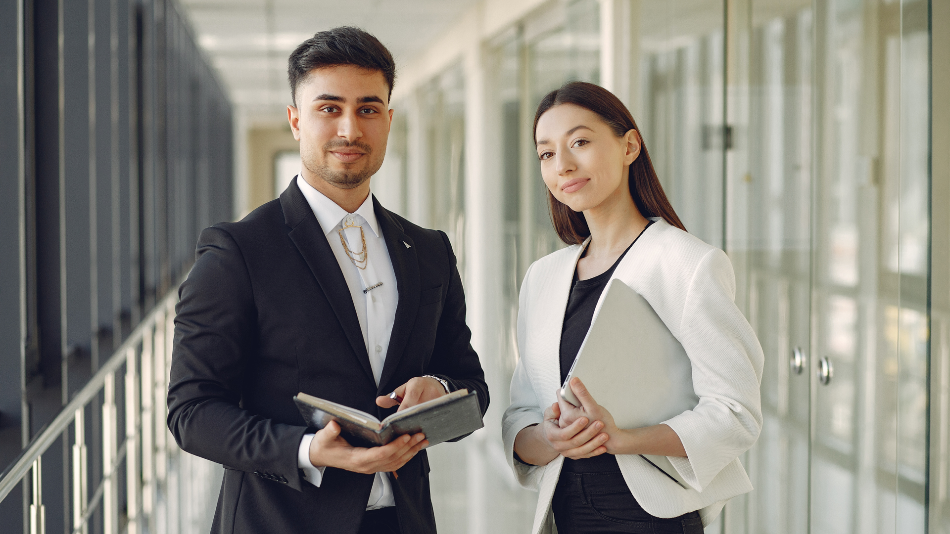 Man and woman in business attire, holding a notebook and laptop, in an office hallway.