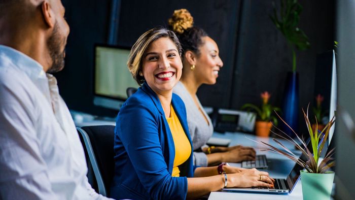 Three coworkers in an office setting. One woman smiles and types on a laptop.
