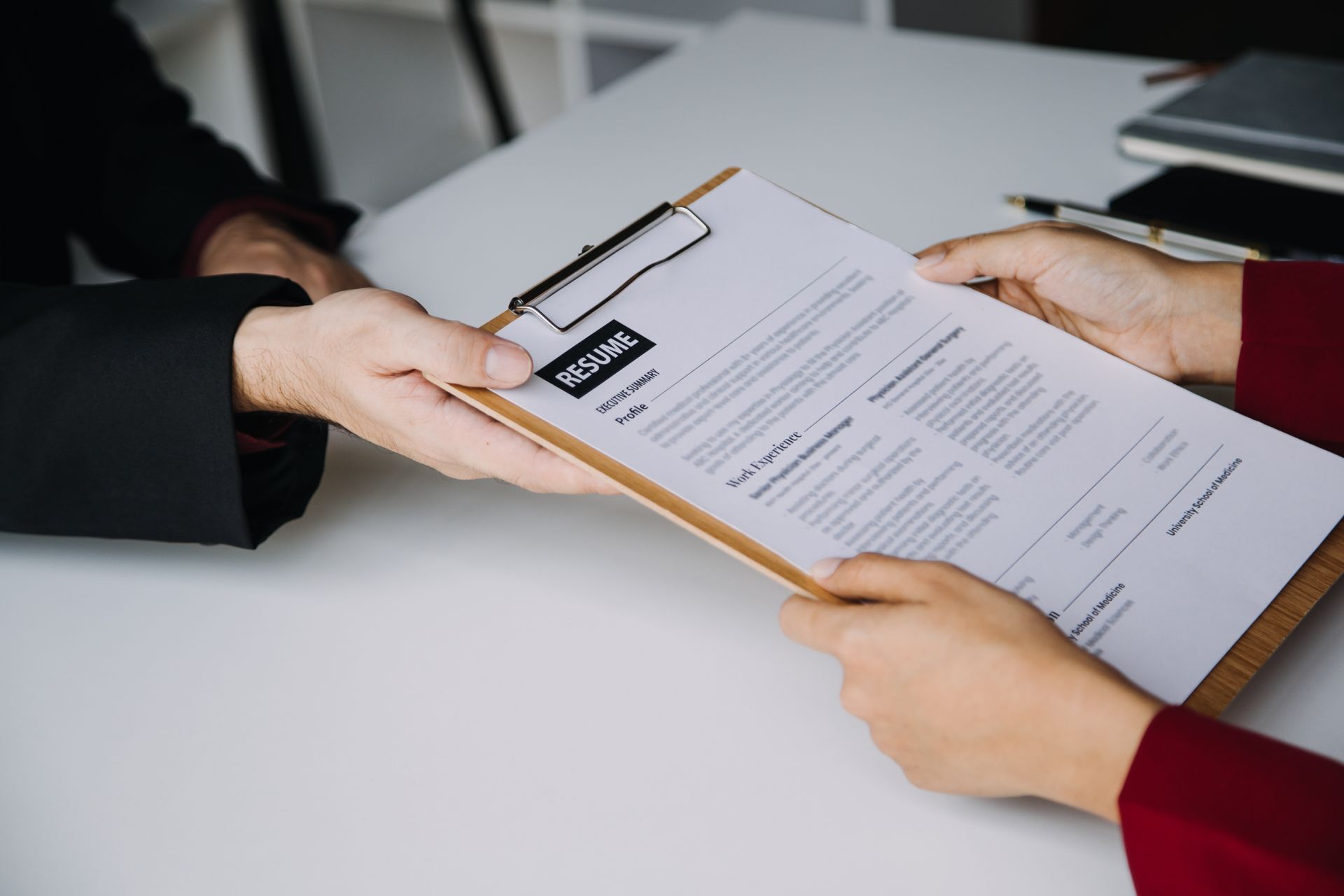 Person handing a resume on a clipboard to another person. White desk.