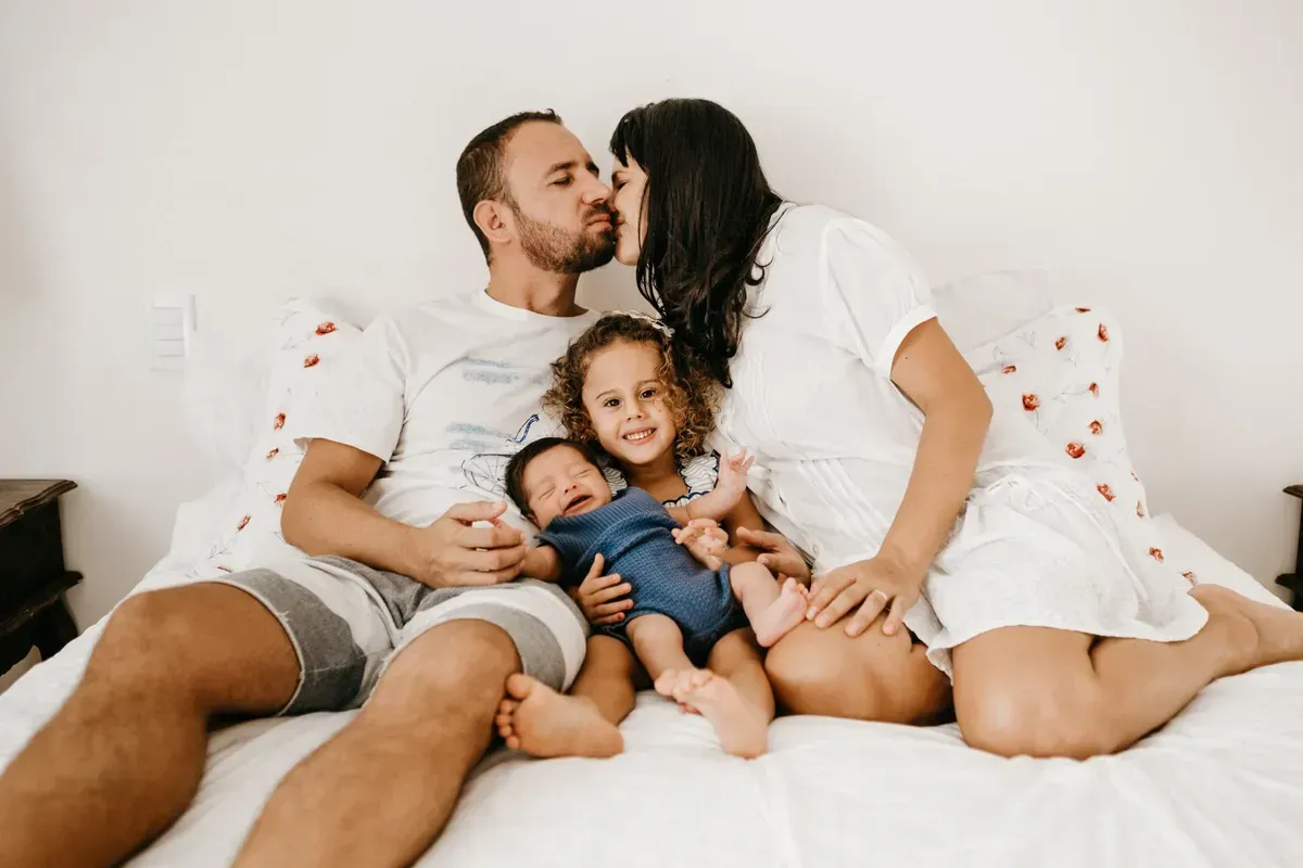 Family of four, parents kissing, with two children on a bed.