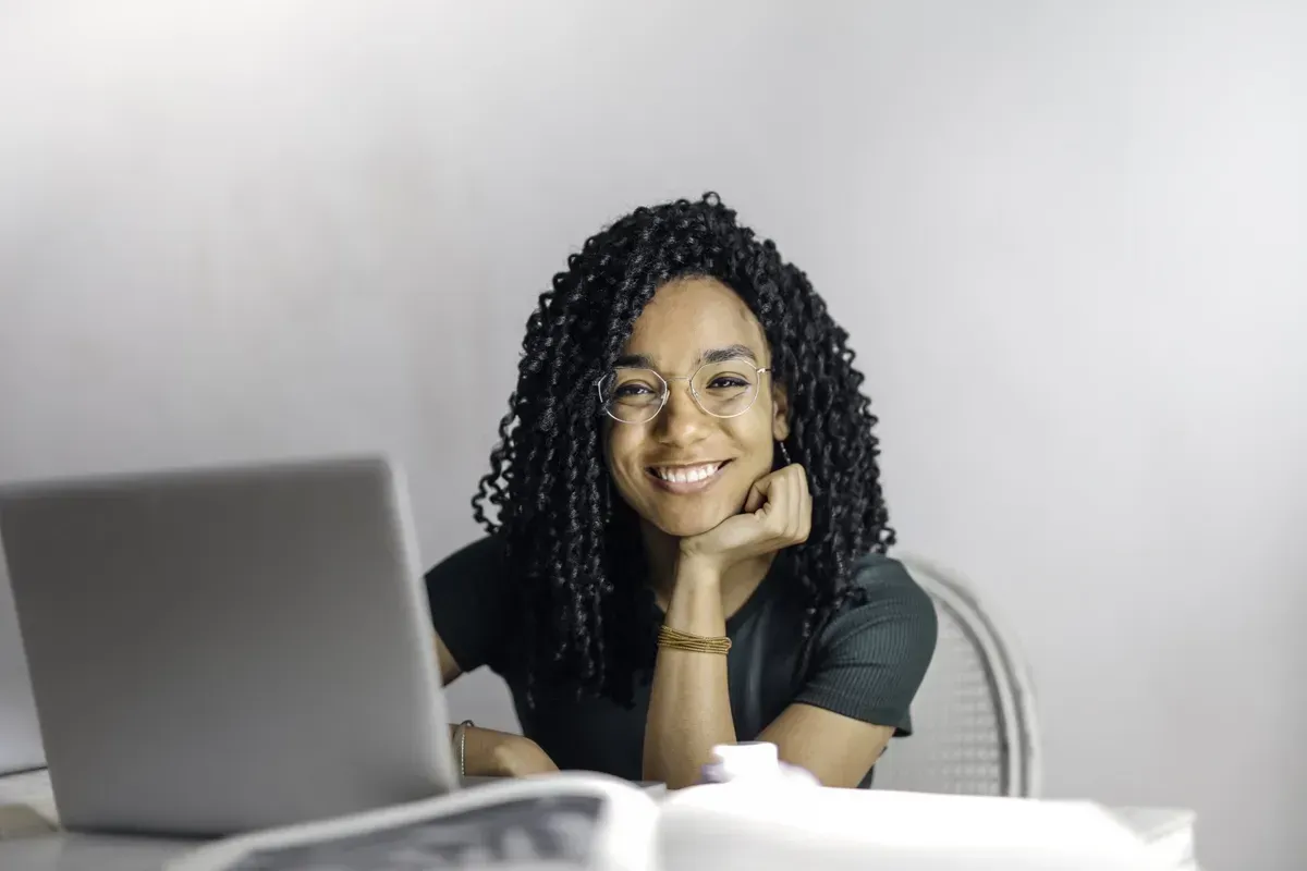Woman with curly dark hair, smiling at laptop, leaning on hand at a desk.