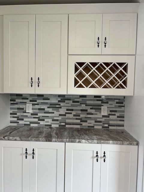 White kitchen cabinets with gray mosaic tile backsplash and dark countertop, featuring a built-in wine rack.
