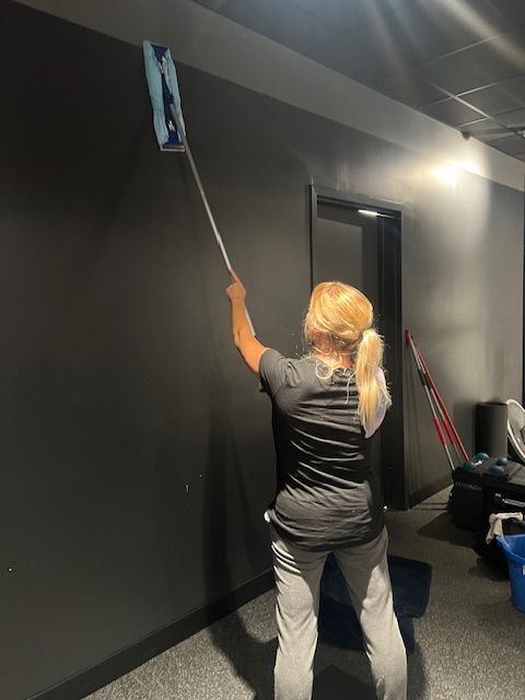 Woman using a long-handled duster to clean a black wall in a gym-like room.