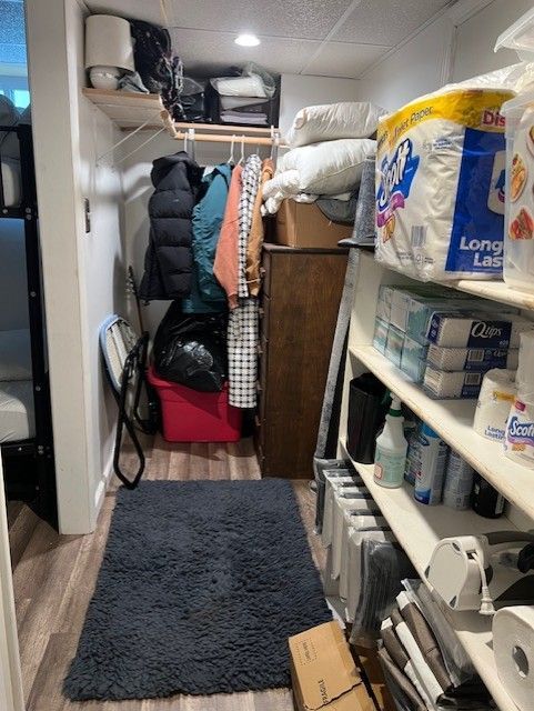 Narrow pantry or closet with shelves of paper goods, hanging coats, and a dark floor mat on the floor.