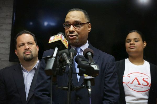 A man in a suit is speaking into a microphone in front of two other people