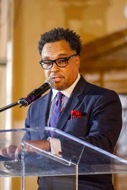 A man in a suit and tie is standing at a podium speaking into a microphone.