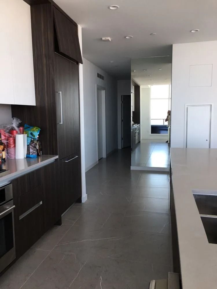Modern kitchen with dark cabinets, gray floor, and a long white island leading to a bright hallway.