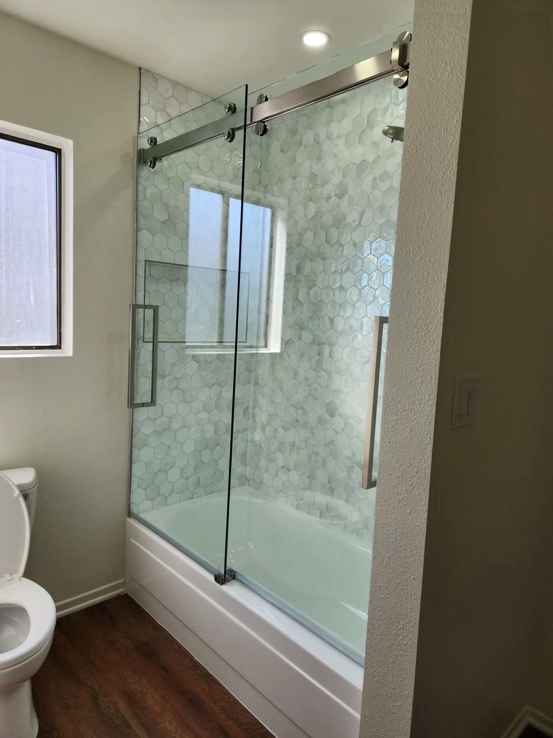 Bathroom with glass sliding shower door, frosted panel, white tub, and wood-look floor.