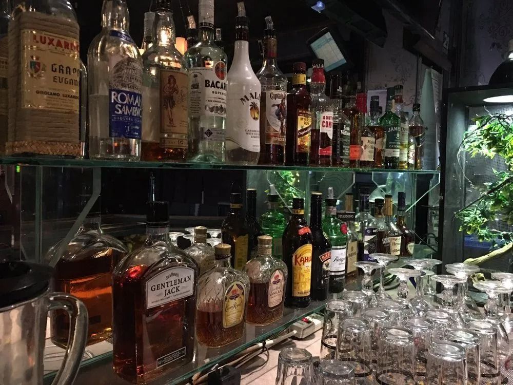 Liquor bottles and glassware on a bar shelf with a leafy window in the background