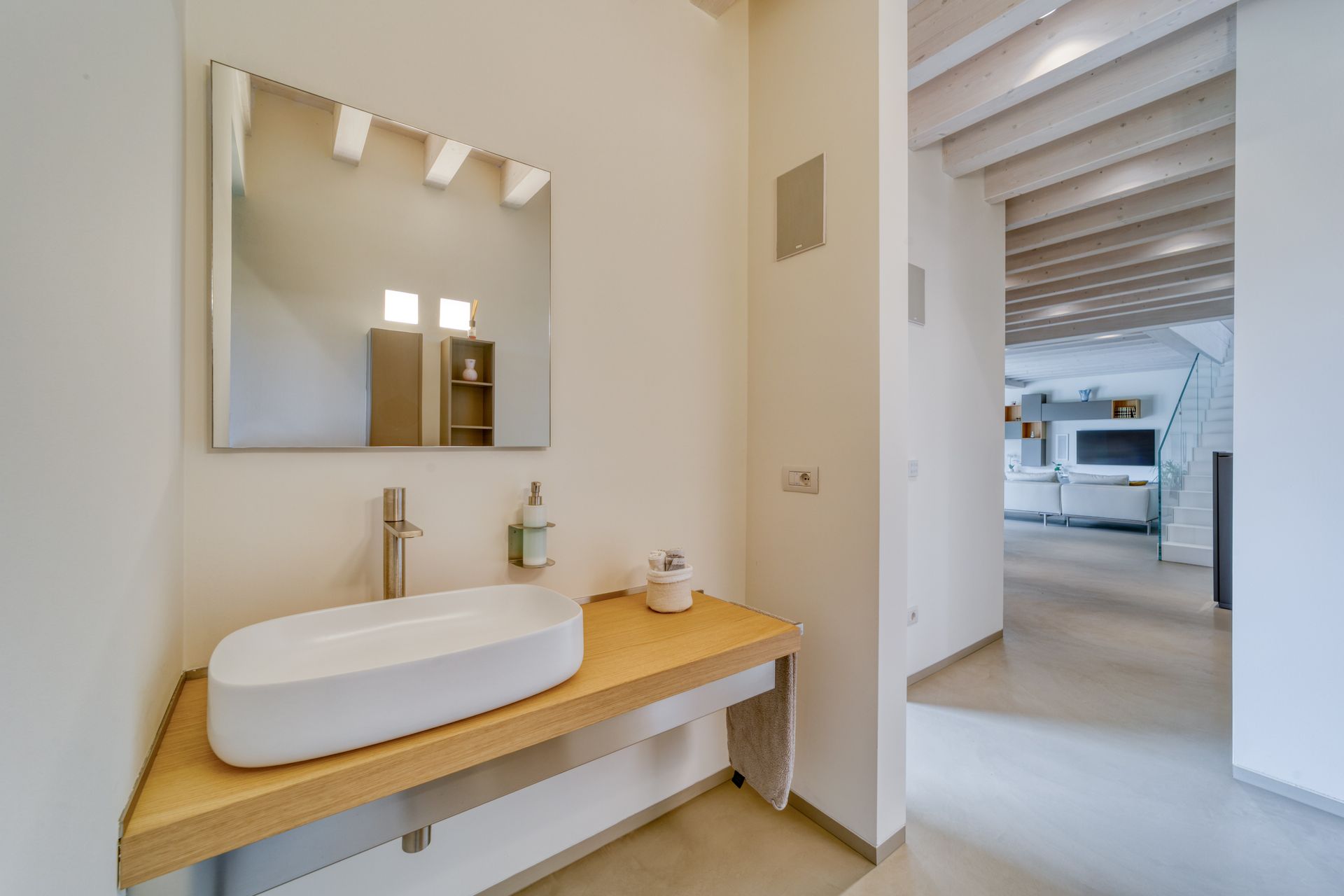 Mobile bagno moderno con lavabo bianco, piano in legno, specchio e porta aperta su una stanza luminosa.