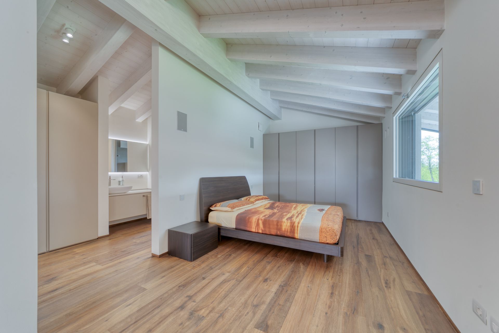 Luminosa camera da letto mansardata con pavimento in legno, letto grigio, pareti bianche e un'ampia finestra.