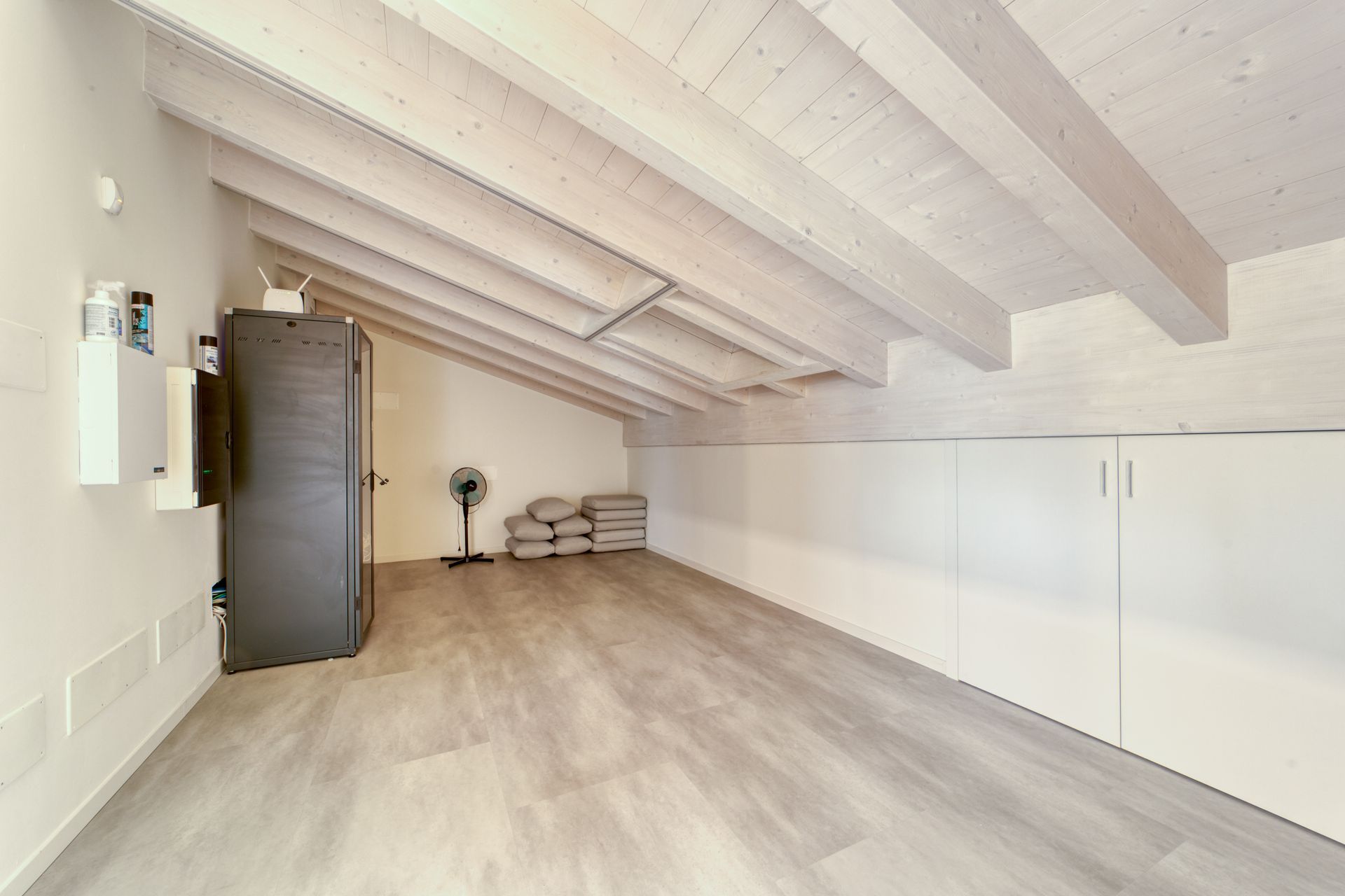 Mansarda vuota con soffitto bianco inclinato, pavimento in legno chiaro e una piccola finestra.