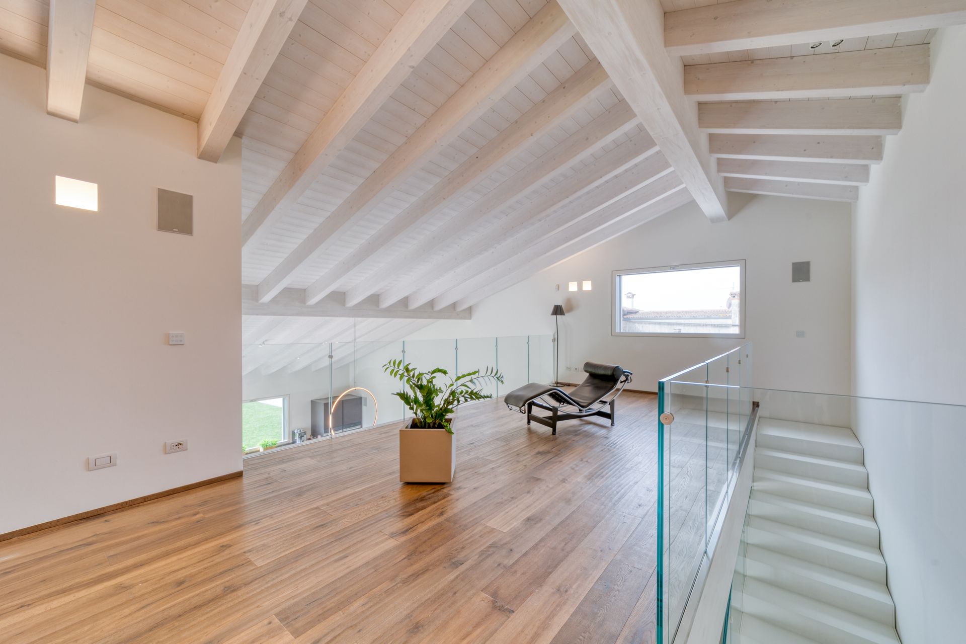 Luminoso interno in stile loft moderno con pareti bianche, pavimento in legno, scala e zona salotto vicino alle grandi finestre.