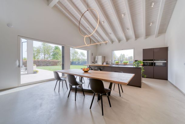 Sala da pranzo moderna e minimalista con lungo tavolo in legno, sedie nere, lampade a sospensione curve e cucina a vista.