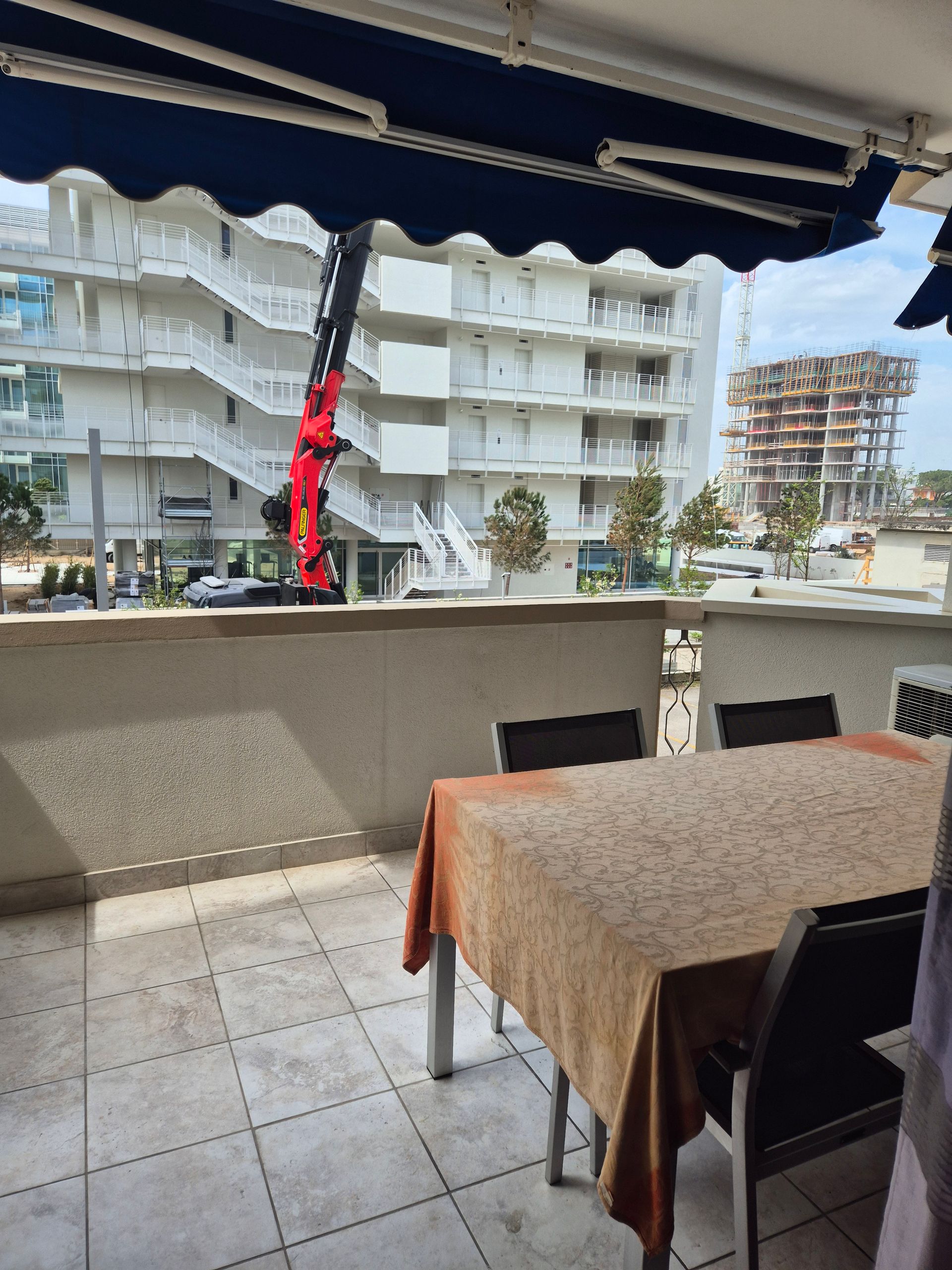 Balcone con tavolo e sedie affacciato su una strada e su palazzi residenziali, sotto una tenda da sole scura.