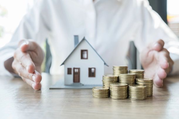 Mani che incorniciano una piccola casa accanto a pile di monete su un tavolo