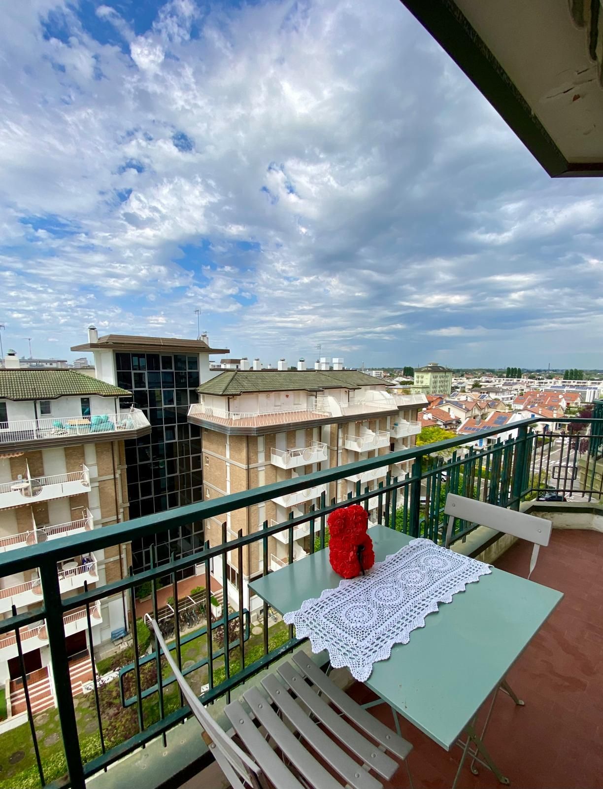 Balcone con tavolino color menta, sedie bianche, vaso rosso e panorama cittadino sotto un cielo nuvoloso.