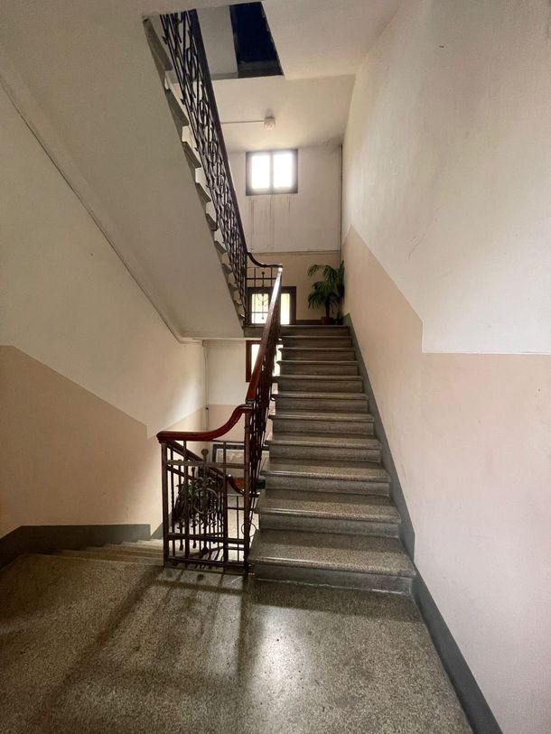 Vano scale interno con pareti beige, gradini in cemento, ringhiera rossa e una piccola finestra sul pianerottolo.