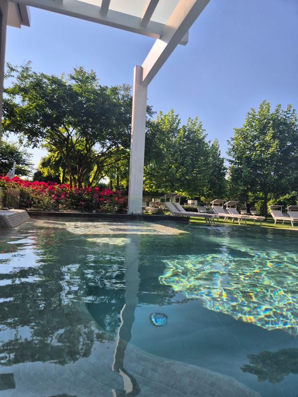 Patio a bordo piscina con acqua azzurra, pergola bianca e alberi verdi sotto un cielo luminoso