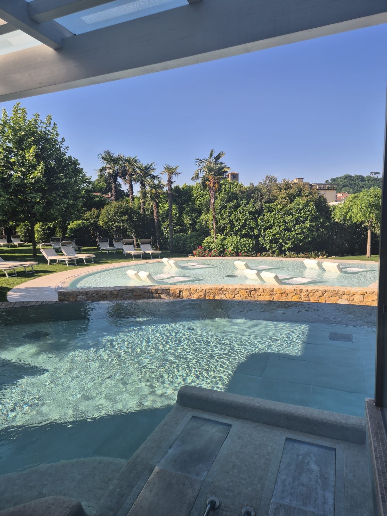 Piscina esterna soleggiata con acqua cristallina, scalini e alberi verdi sullo sfondo.
