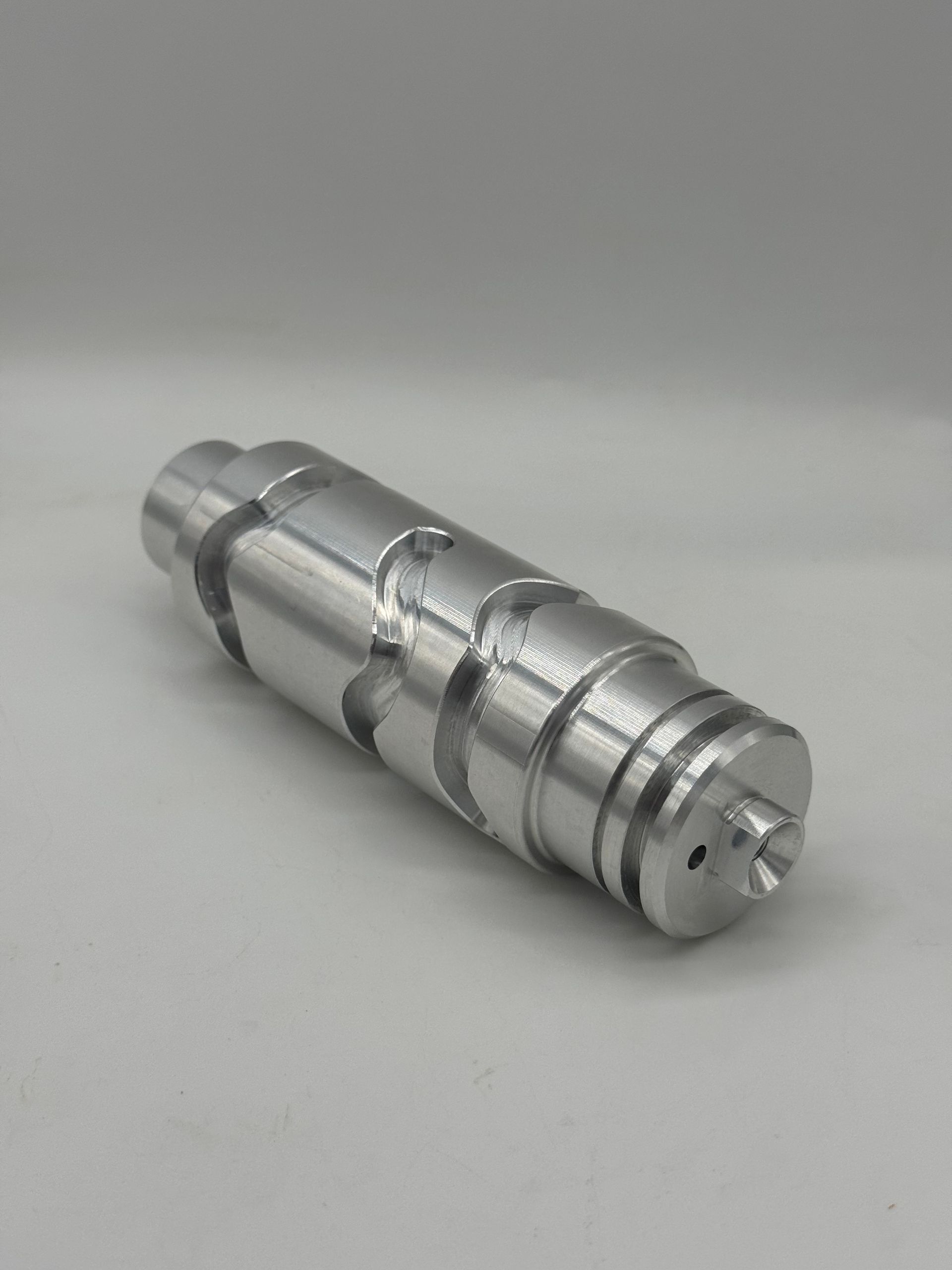 A machined cylindrical silver metal part with spiraling grooves and a small hole on the flat end, set against a gray background.