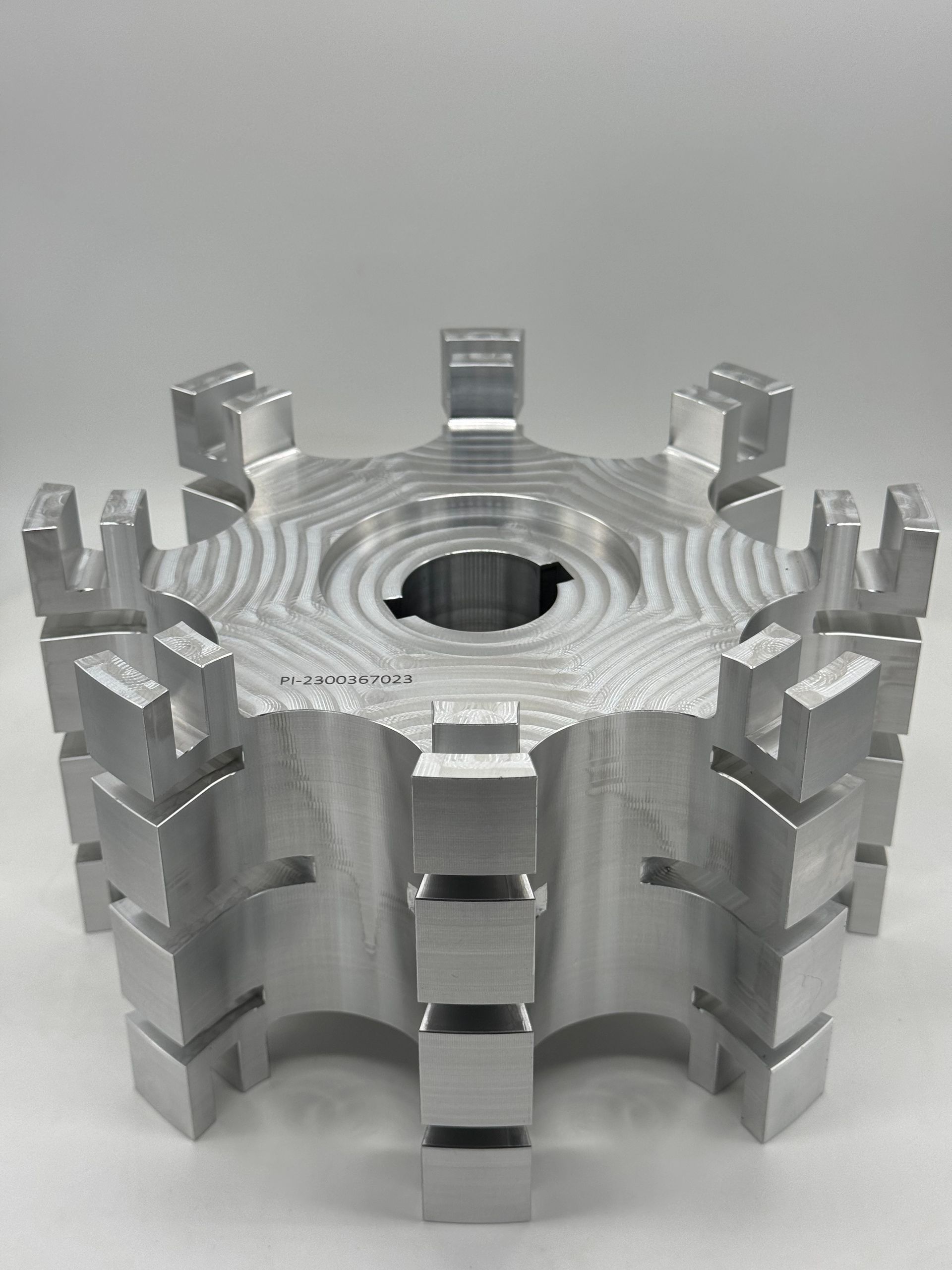 A silver, machined metal rotor or hub with eight evenly spaced, notched vertical fins around its circular core.