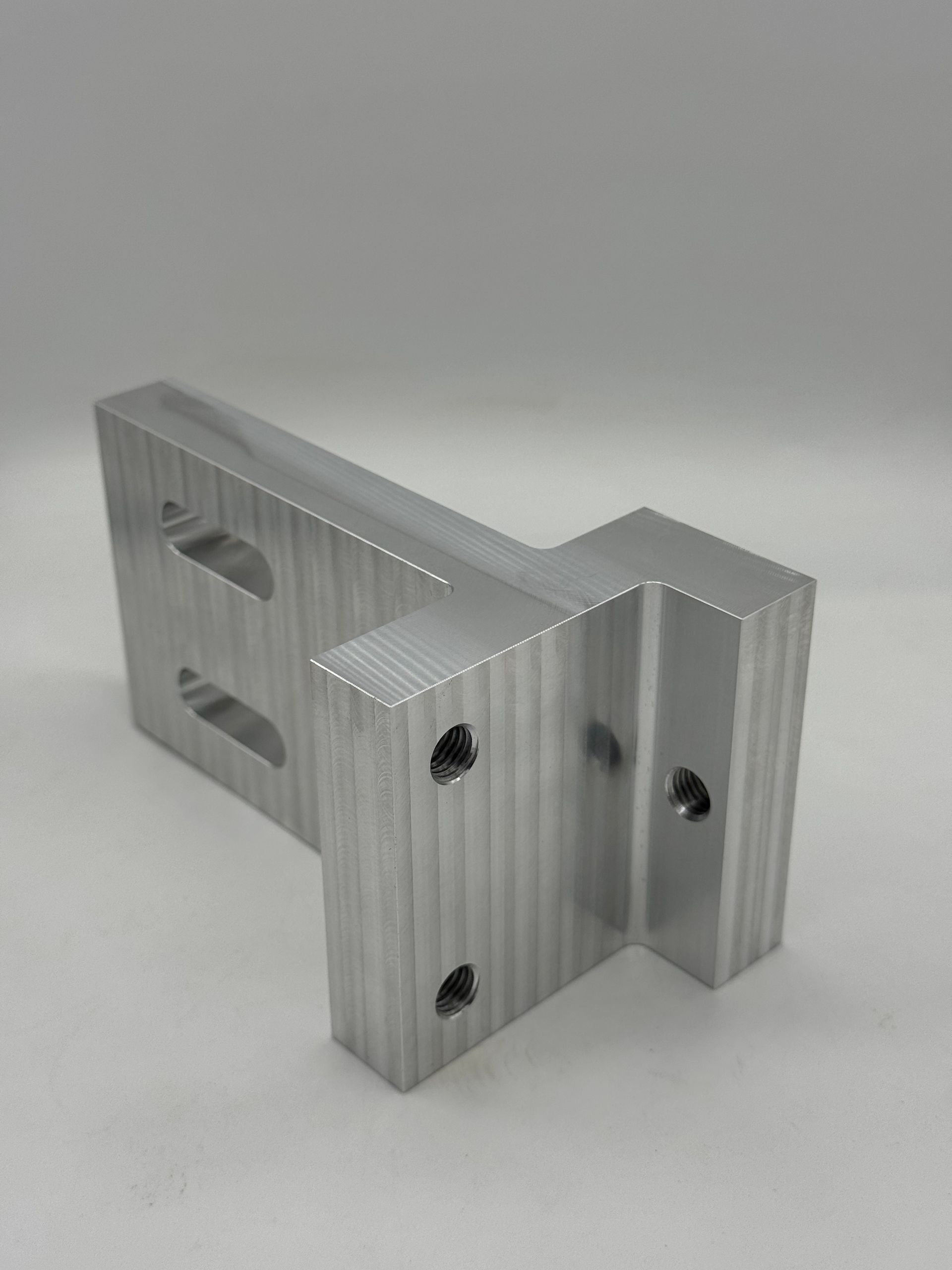 A machined aluminum L-bracket with two slotted holes on one side and three circular holes on the other.