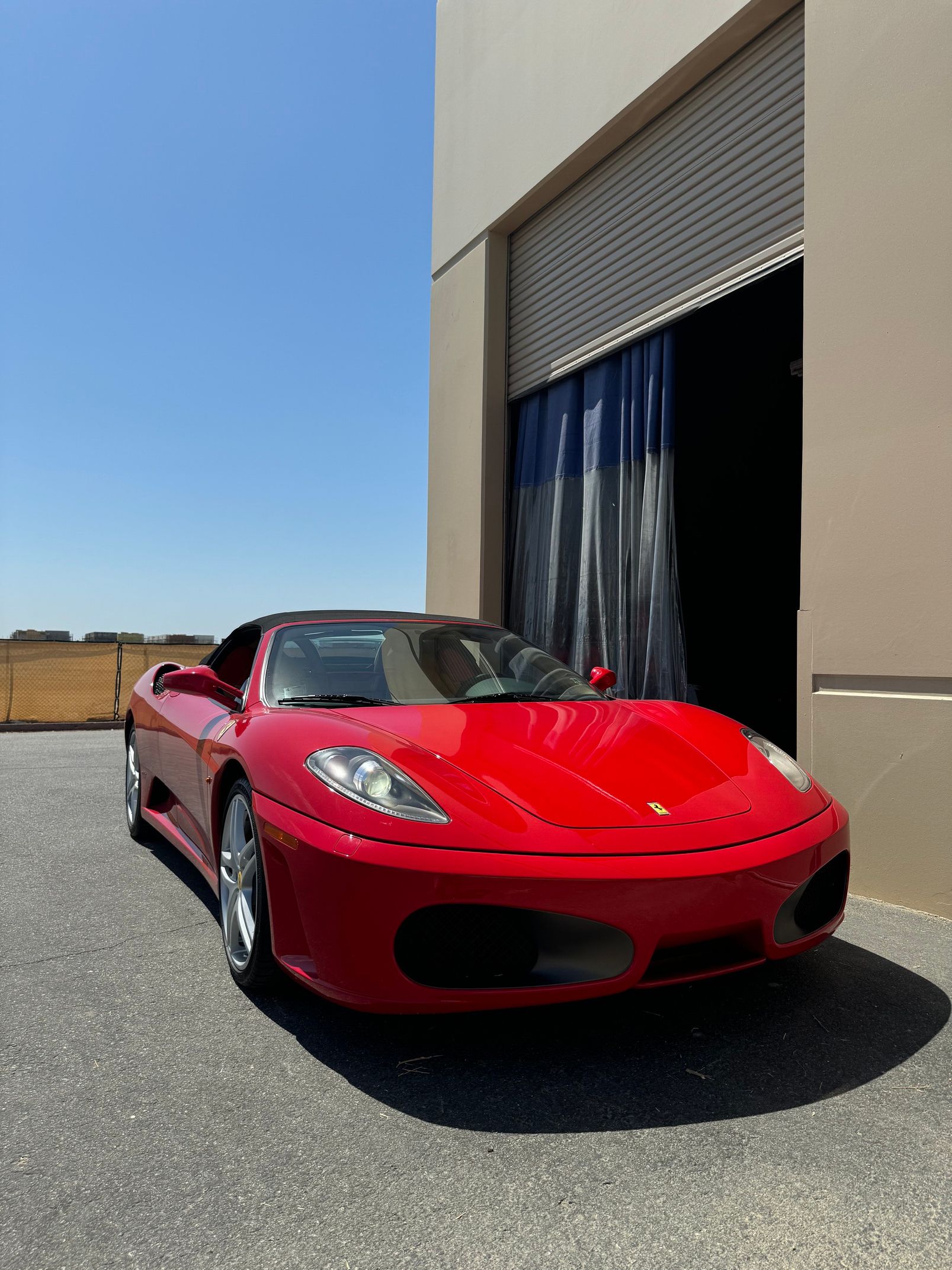 Red sports car parked outside a beige building with an open garage door and blue sky.