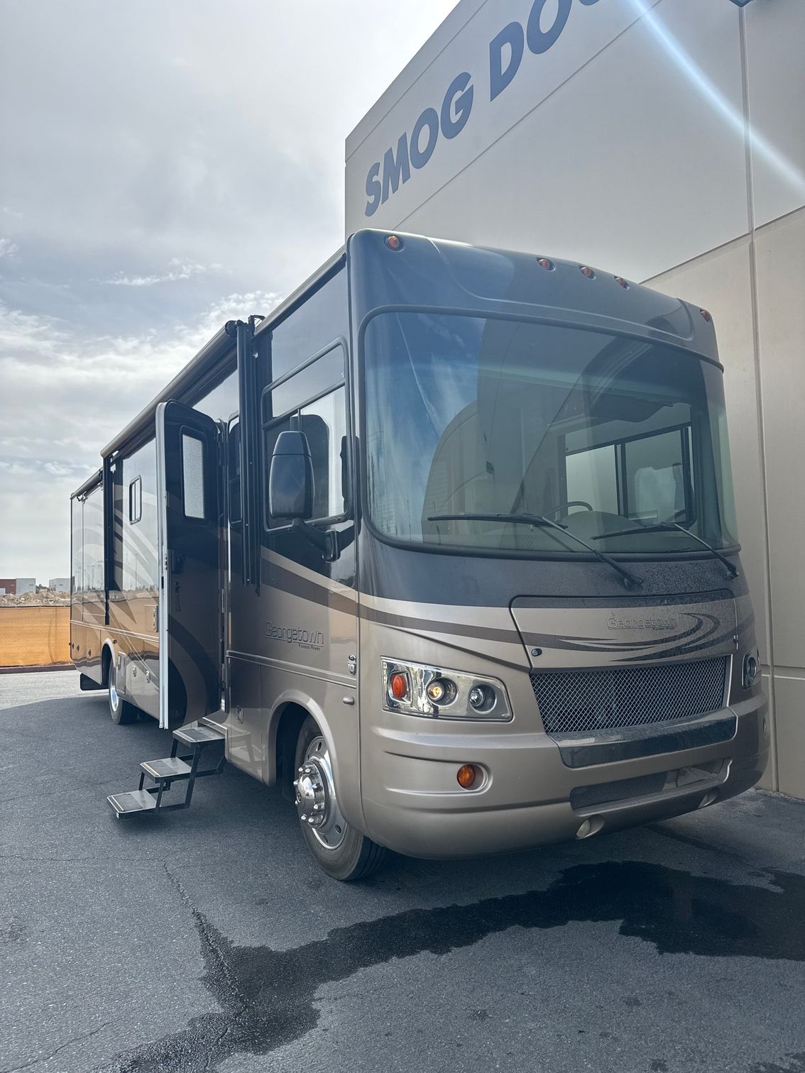 Silver motorhome with slide-outs parked outside a building on a cloudy day