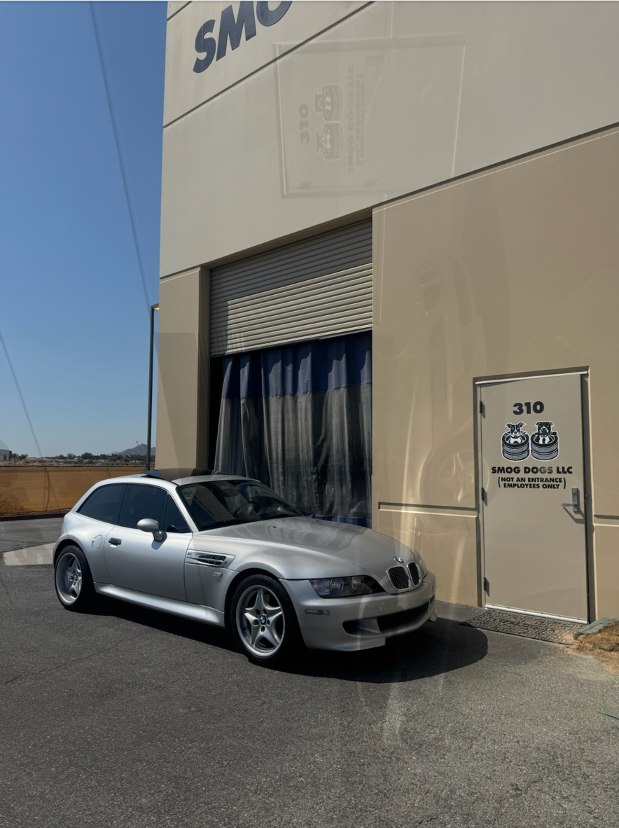 Silver sports coupe parked outside a beige building with a garage door and signage.