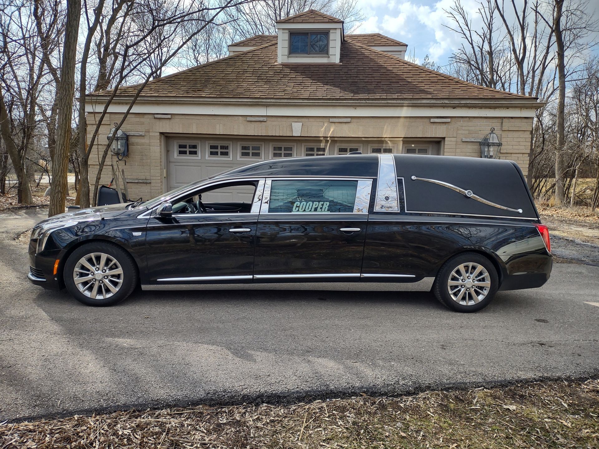 Black hearse parked on a suburban street in front of a beige house and bare trees.