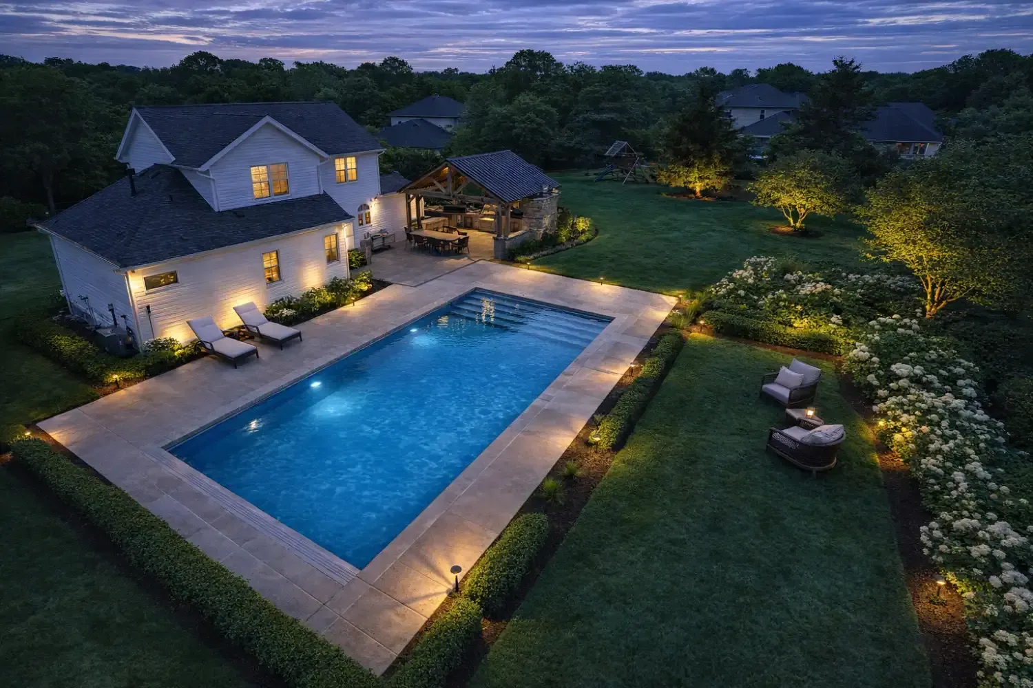 An aerial twilight view of a white house with a lit swimming pool, stone patio, and landscaped yard.