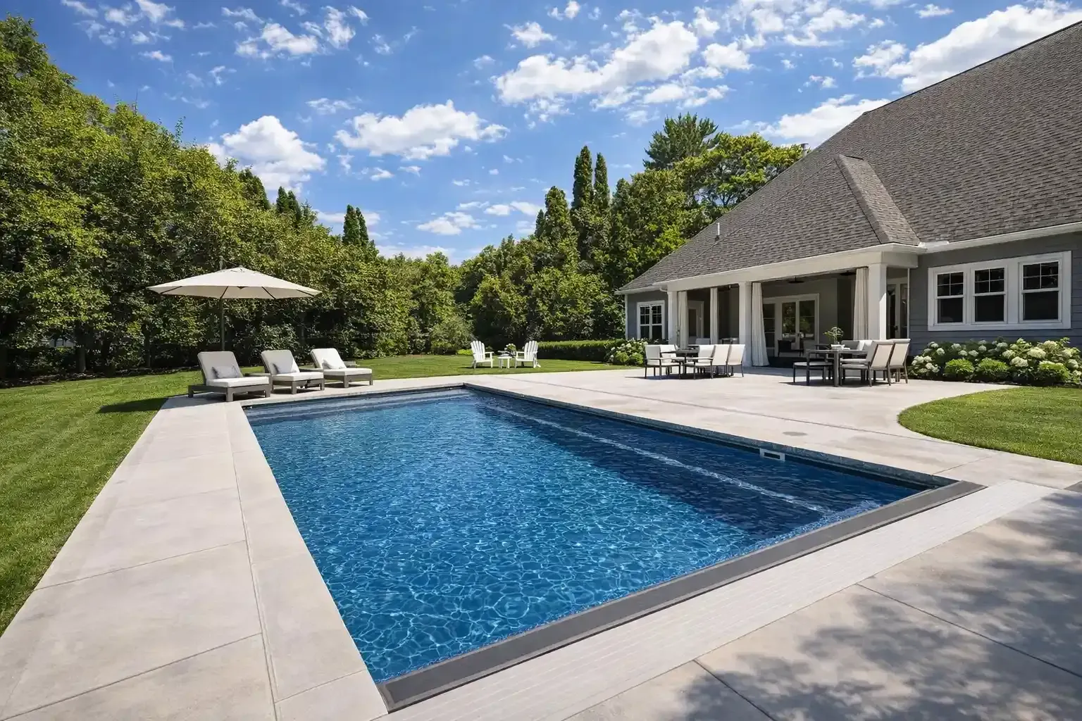 A rectangular swimming pool with deep blue water sits on a stone patio next to a house under a sunny, blue sky.
