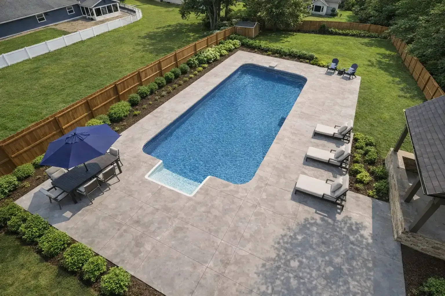 A rectangular swimming pool on a stone patio with lounge chairs, an umbrella, and a wooden fence in a backyard setting.