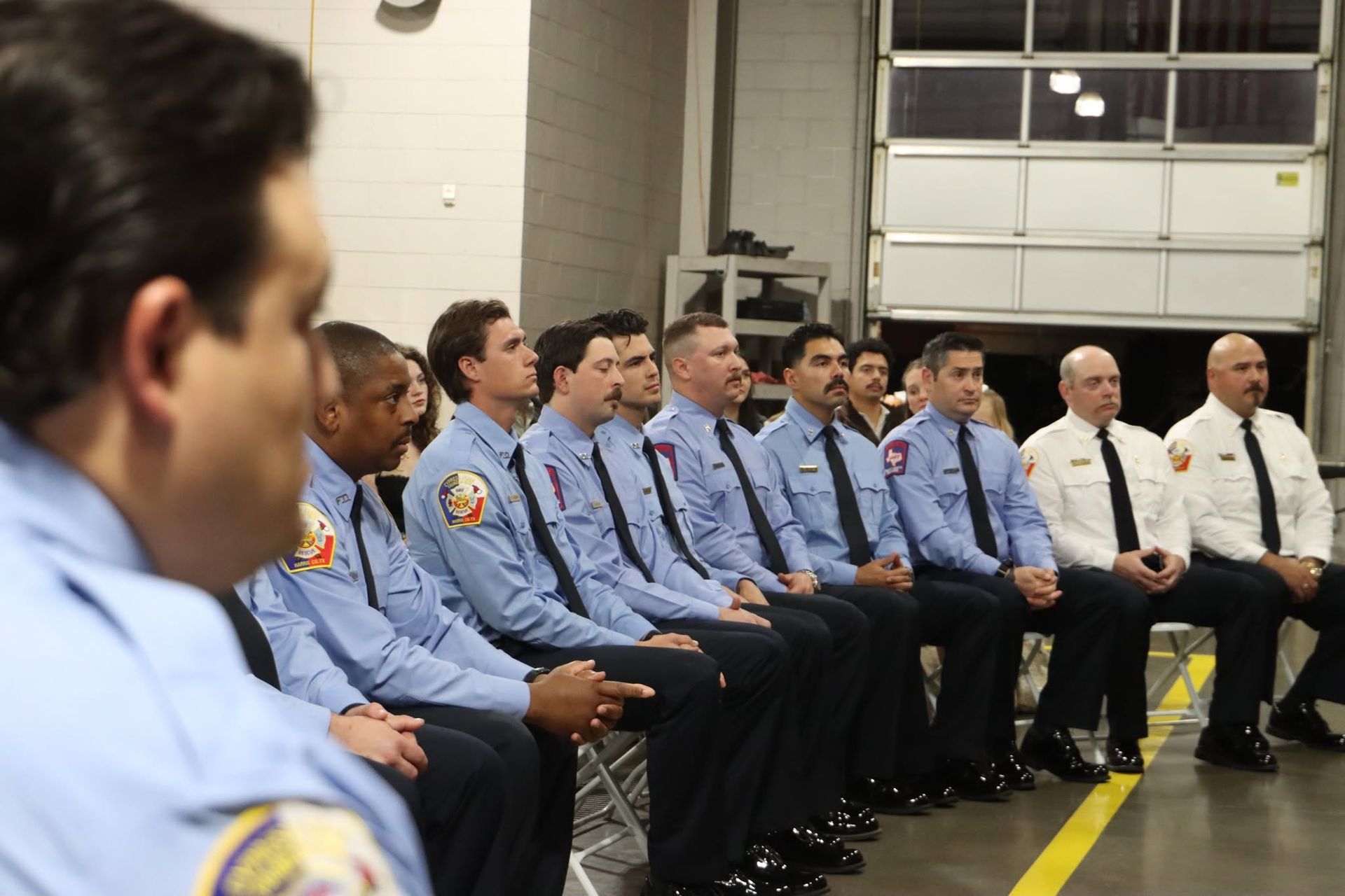 Uniformed officers seated in rows during a briefing inside a garage or training bay