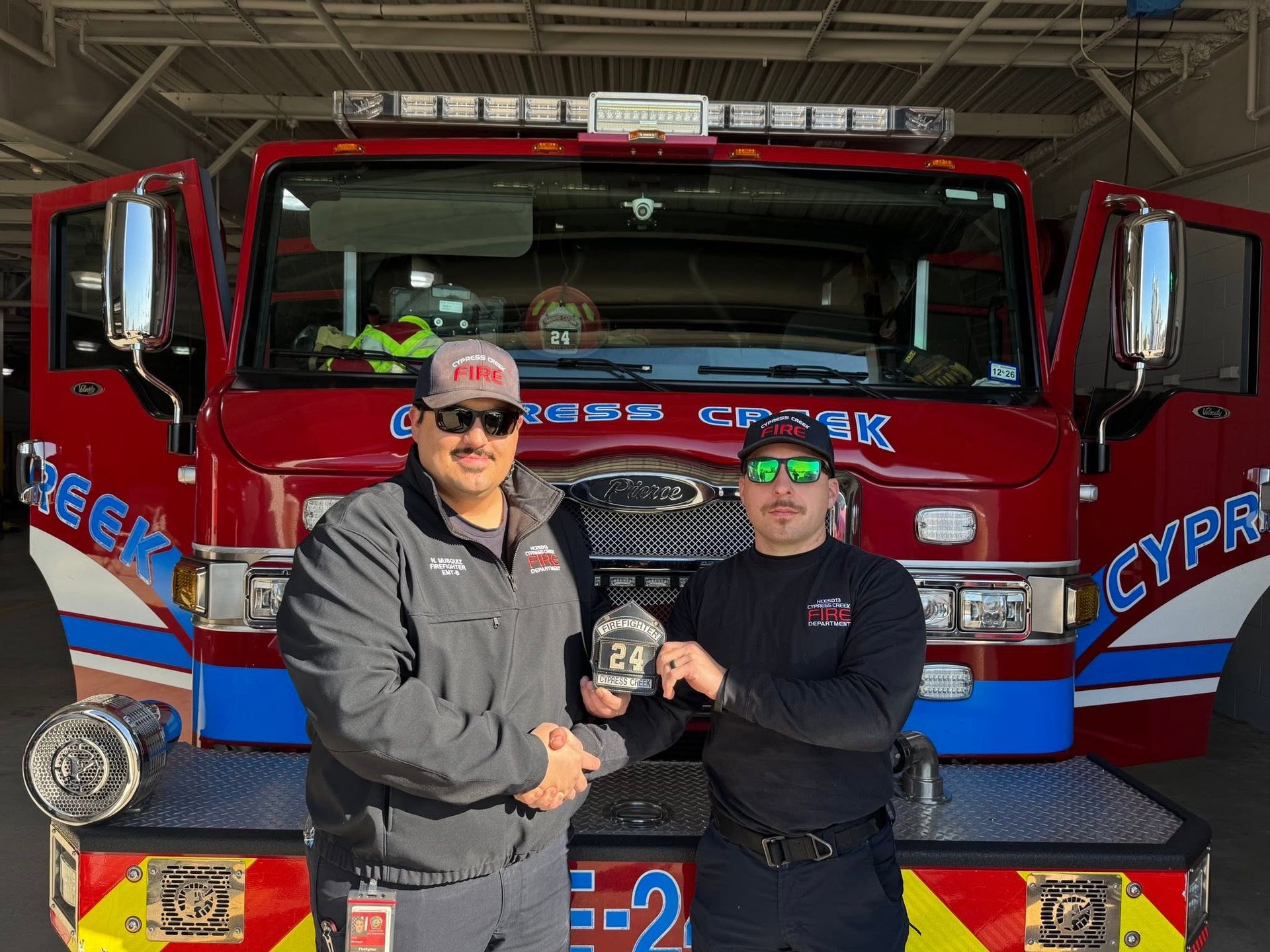 Two firefighters holding a small plaque in front of a red fire truck at the station