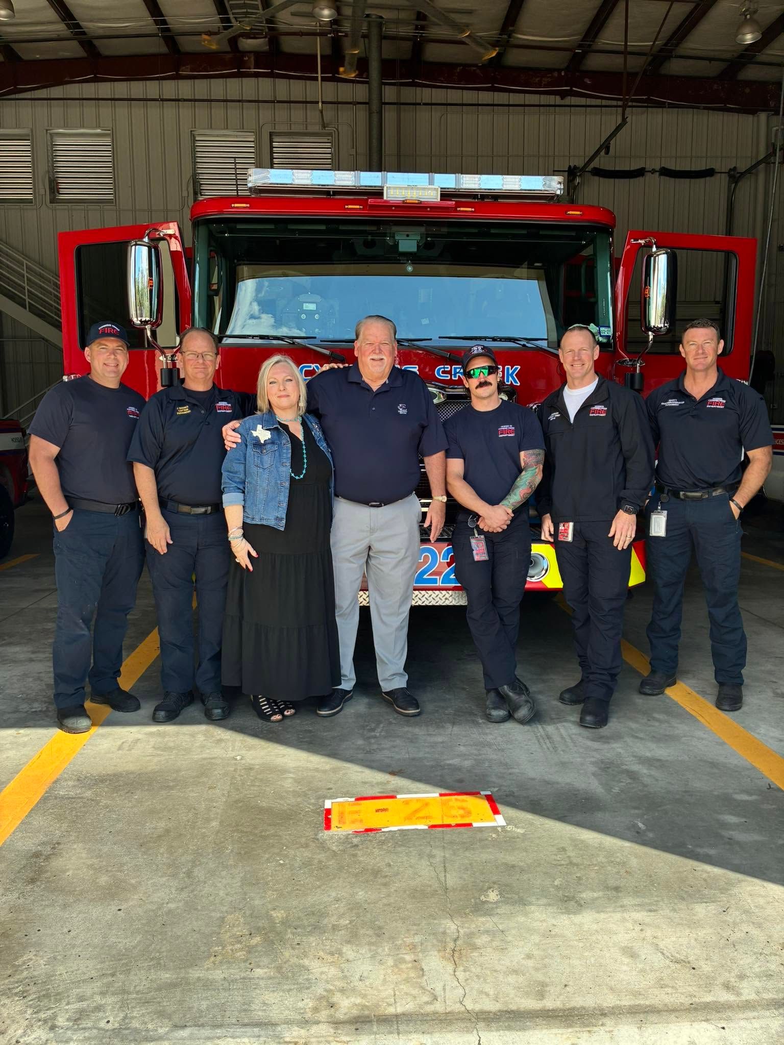 Six firefighters standing in front of a red fire truck inside a fire station.