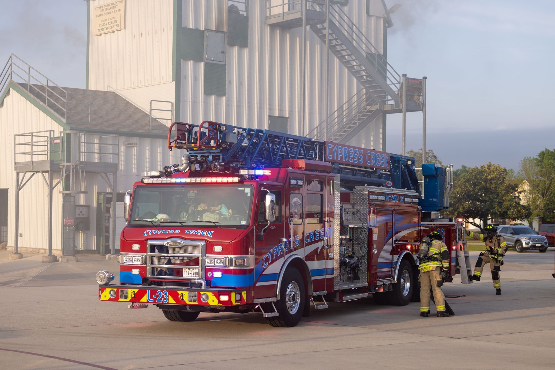 Red fire ladder truck with flashing lights parked by a building, firefighters standing nearby.