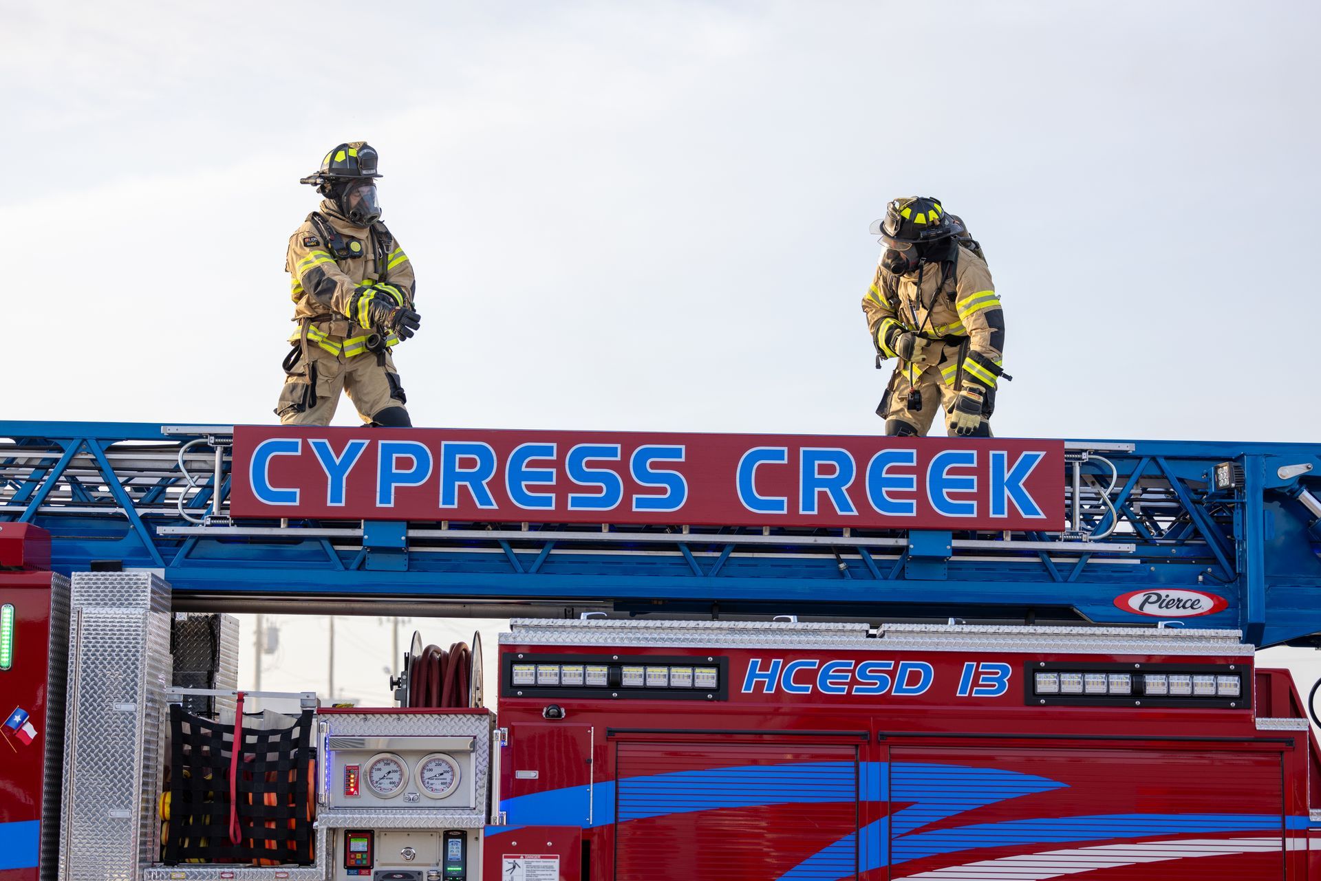 Firefighters on a red fire truck ladder with “CYPRESS CREEK” sign against the sky