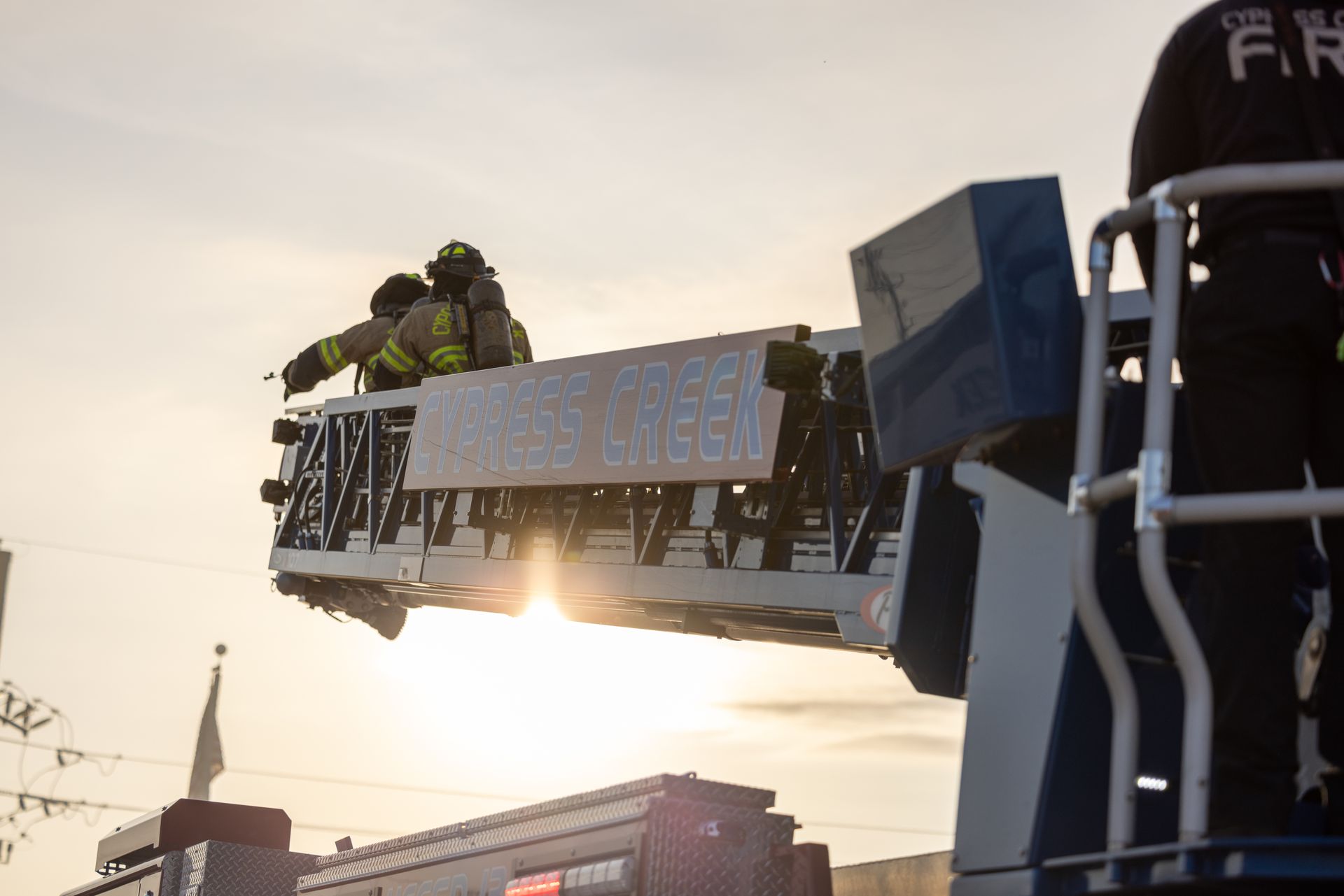 Firefighters on an aerial ladder hoist a banner at sunset.