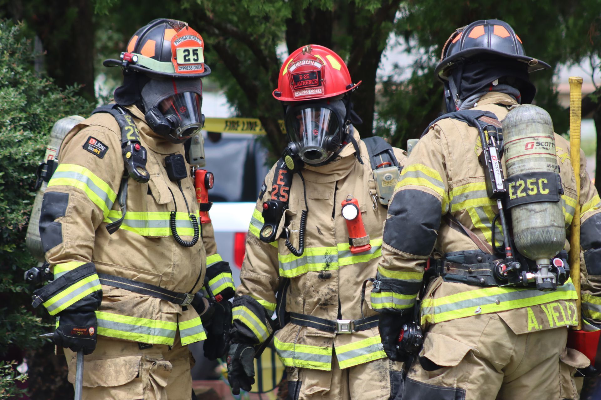 Three firefighters in protective gear standing outdoors, one in a red helmet centered between two in black helmets.