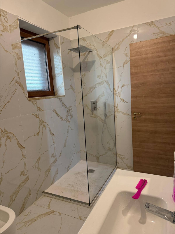 Bagno moderno con box doccia in vetro, piastrelle effetto marmo, finestra e lavabo bianco con maniglia rosa.