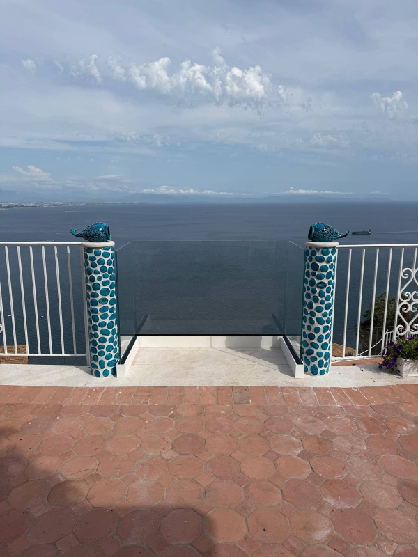 Vista sull'oceano da un patio piastrellato con ringhiere bianche e colonne a mosaico blu sotto un cielo nuvoloso.
