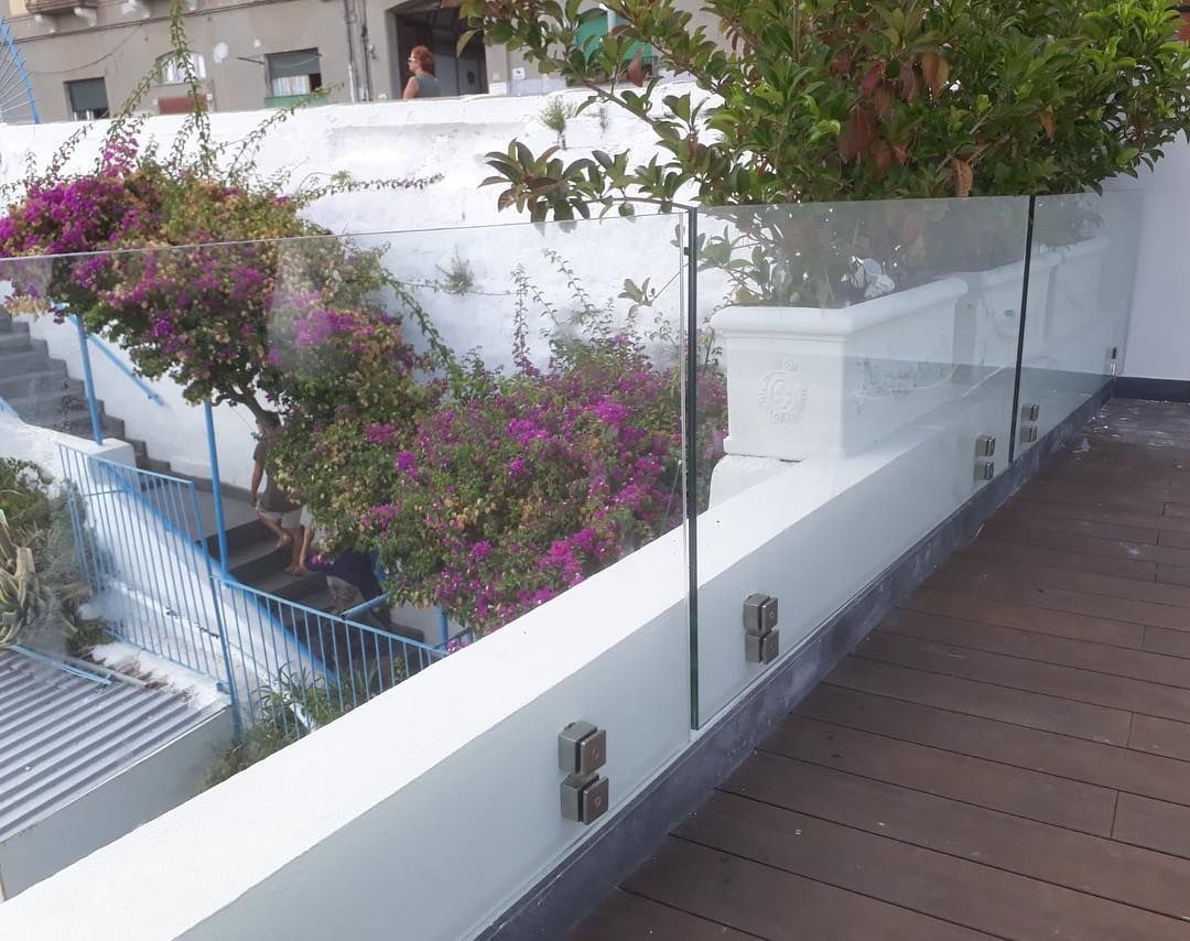 Ringhiera in vetro del balcone accanto a una terrazza in legno, con pareti bianche e arbusti fioriti rosa sullo sfondo.